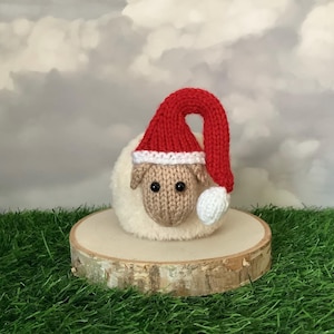 Könnte beinhalten: Eine gestrickte Schaf-Figur mit roter Weihnachtsmütze mit weißem Rand und Bommel. Das Schaf hat ein beiges Gesicht und steht auf einer Holzscheibe, mit grünem Gras und einem bewölkten Himmel im Hintergrund.