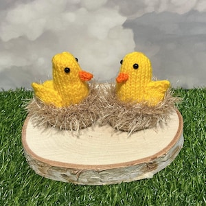 Puede incluir: Dos patitos amarillos tejidos a mano con picos naranjas y ojos negros, anidados en nidos de hilo marrón. Los patitos están sobre una rodaja de madera redonda, ideal para la decoración de Pascua o una guardería.