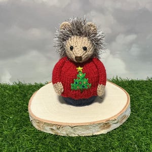Peut inclure: Figurine de hérisson tricoté à la main, vêtu d'un pull rouge orné d'un sapin de Noël vert. Le hérisson a un visage beige et un dos épineux argenté et marron. Il est présenté sur une tranche de bois, sur fond d'herbe verte et de ciel nuageux.