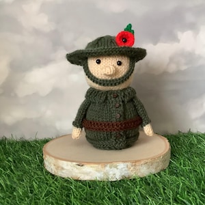 May include: A knitted doll wearing a green hat with a red poppy, a green jacket, and a brown belt. The doll has a beige face and a knitted beard. It is displayed on a wooden slice against a cloudy backdrop.