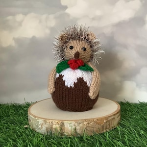 以下が含まれることがあります： 茶色の体、ベージュの腕、とげのある頭を持つ、編み物のハリネズミの置物。 ハリネズミは緑と赤のヒイラギの飾りをしています。 小さな木の輪切りに飾られています。