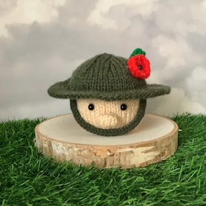 May include: A knitted figure wearing a green hat adorned with a red poppy. The figure has a beige face and black eyes. It is presented on a wooden slice, set against a backdrop of grass and a cloudy sky. The hat features a brim.