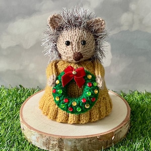 May include: A knitted hedgehog figurine with a tan face, black eyes, and spiky dark gray and white fur. The hedgehog wears a mustard yellow sweater and holds a small green wreath decorated with red and yellow sequins and a red velvet bow.