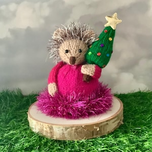 May include: A knitted hedgehog figure in a pink dress, holding a green Christmas tree with colourful baubles and a gold star. It stands on a wooden slice base with a pink tinsel trim, perfect for festive decor.