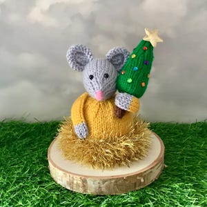 Peut inclure: Figurine de souris tricotée à la main, vêtue d'un pull jaune, tenant un sapin de Noël vert orné de décorations colorées et d'une étoile dorée. La souris grise a un nez rose et est posée sur une base en bois avec des guirlandes dorées.