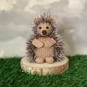 以下が含まれることがあります： 手編みのハリネズミの置物。茶色と銀色のキラキラした飾り毛が特徴です。ハリネズミはベージュ色の体、小さな耳、茶色の鼻と目を持っています。小さな丸い木の輪切りに飾られており、インテリアに最適です。