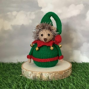May include: A knitted hedgehog ornament in an elf costume. The hedgehog has a beige face and silver tinsel 'hair', wearing a green outfit with red trim and gold bells. It sits on a wooden slice, set against a cloudy sky background.