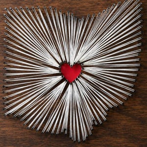 May include: A string art piece on a dark brown wooden board featuring a map of Ohio outlined with silver nails and white thread. The threads radiate from the perimeter toward a central red heart shape positioned in the middle of the state map.