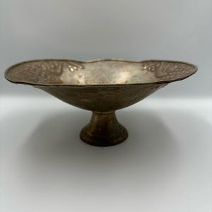 May include: A vintage, pedestal-style bowl with a wide, scalloped rim. The bowl has a metallic finish with a textured surface and a decorative pattern around the rim. The base is a short, rounded pedestal.