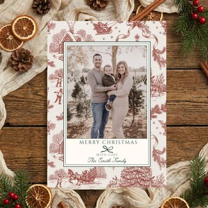 Pode incluir: Um cartão de Natal com uma fotografia de família emoldurada em branco. O cartão tem um padrão toile vermelho e branco com cenas de inverno. O texto "MERRY CHRISTMAS WITH LOVE The Smith Family" é exibido abaixo da foto.