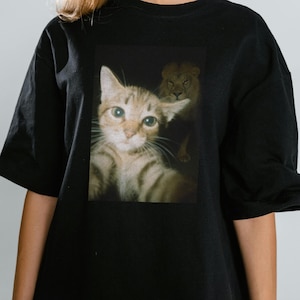 May include: Black t-shirt featuring a photographic print of a tabby cat taking a selfie, with a lion subtly visible in the background. The cat has green eyes and is looking directly at the viewer. The lion is in the background.