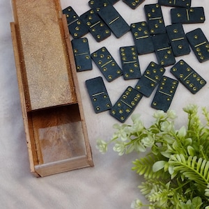 May include: A set of dominoes with a wooden box. The dominoes are dark teal with gold dots. The box is rectangular, with a clear plastic sliding lid. Green and white floral accents are in the background.