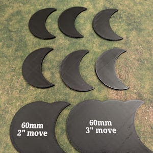 K&ouml;nnte beinhalten: Eine Sammlung schwarzer, halbmondf&ouml;rmiger Spielmarken. Zwei gr&ouml;&szlig;ere Marken sind mit "60mm 2" move" und "60mm 3" move" beschriftet. Die Marken sind auf einer gr&uuml;n-braunen Oberfl&auml;che angeordnet.