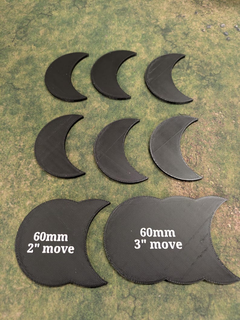 K&ouml;nnte beinhalten: Eine Sammlung schwarzer, halbmondf&ouml;rmiger Spielmarken. Zwei gr&ouml;&szlig;ere Marken sind mit "60mm 2" move" und "60mm 3" move" beschriftet. Die Marken sind auf einer gr&uuml;n-braunen Oberfl&auml;che angeordnet.