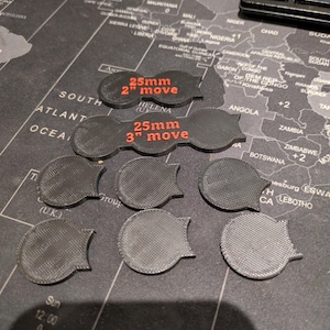 K&ouml;nnte beinhalten: Eine Sammlung schwarzer, 3D-gedruckter Spielmarken. Zwei Marken tragen den roten Text "25mm 2" move" und "25mm 3" move". Die anderen Marken sind kreisf&ouml;rmig mit strukturierter Oberfl&auml;che und gebogenem Rand.
