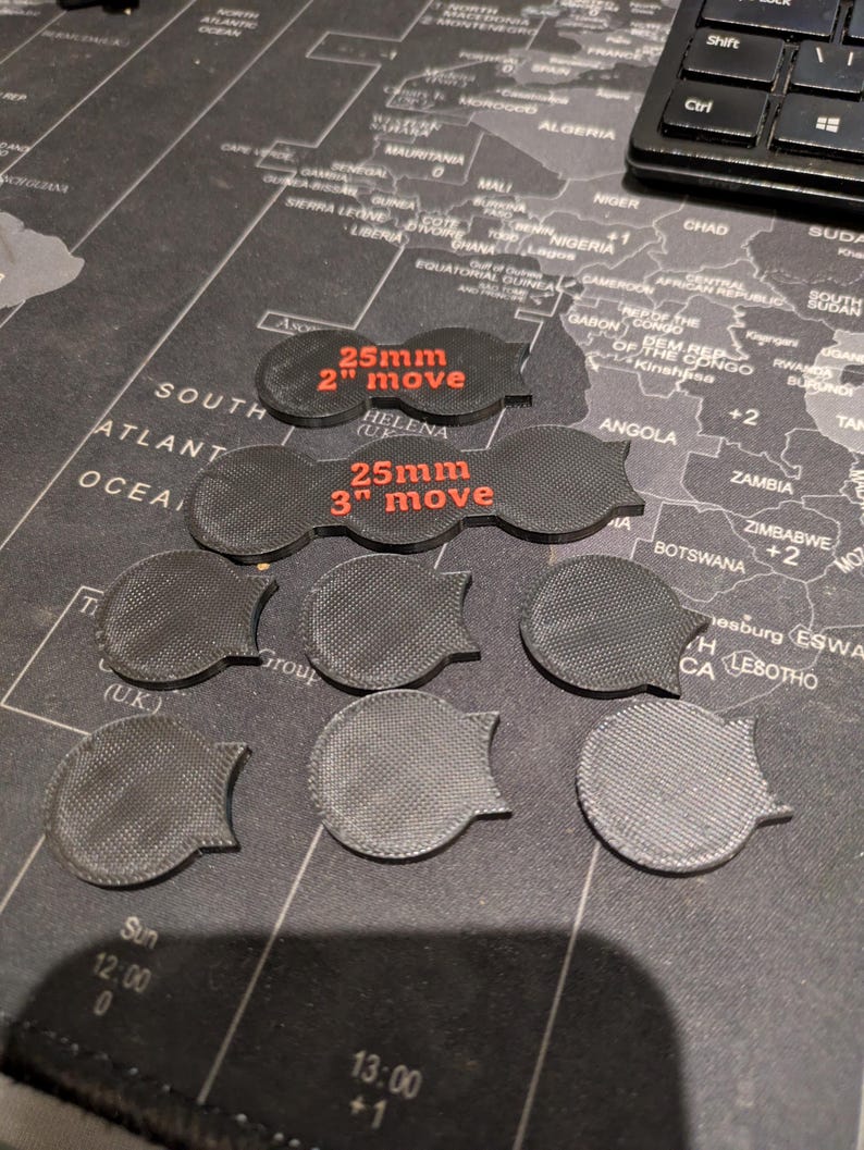 K&ouml;nnte beinhalten: Eine Sammlung schwarzer, 3D-gedruckter Spielmarken. Zwei Marken tragen den roten Text "25mm 2" move" und "25mm 3" move". Die anderen Marken sind kreisf&ouml;rmig mit strukturierter Oberfl&auml;che und gebogenem Rand.