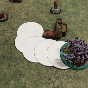 K&ouml;nnte beinhalten: Eine Tabletop-Spielszene mit einer lila Monster-Miniatur, wei&szlig;en kreisf&ouml;rmigen Spielsteinen und Miniaturgeb&auml;uden auf einer gr&uuml;nen Oberfl&auml;che. Das Monster hat mehrere augen&auml;hnliche Merkmale.