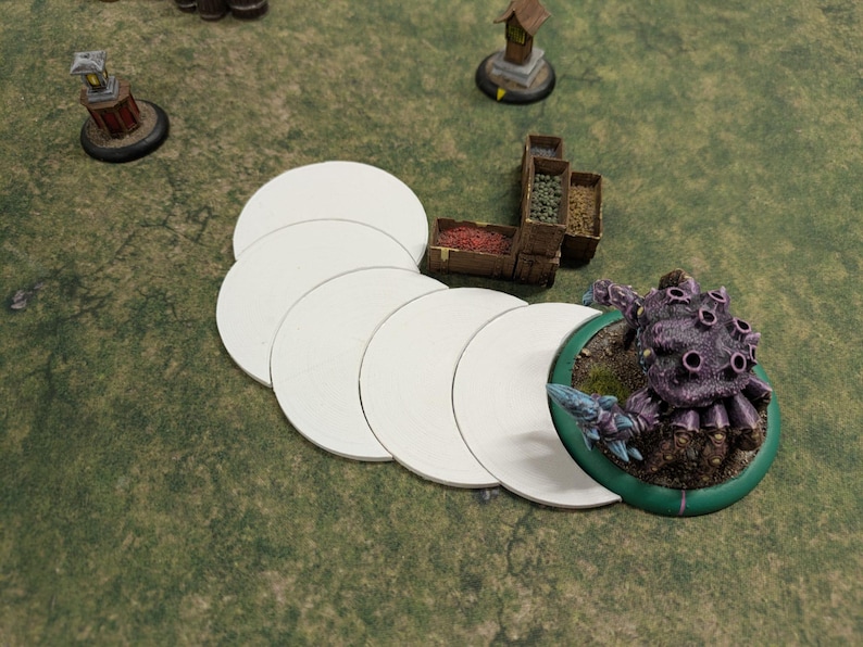 K&ouml;nnte beinhalten: Eine Tabletop-Spielszene mit einer lila Monster-Miniatur, wei&szlig;en kreisf&ouml;rmigen Spielsteinen und Miniaturgeb&auml;uden auf einer gr&uuml;nen Oberfl&auml;che. Das Monster hat mehrere augen&auml;hnliche Merkmale.