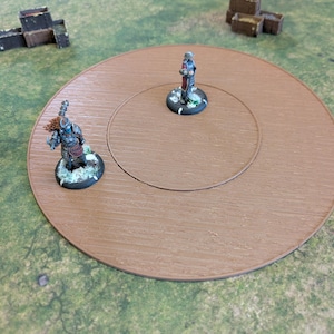Könnte beinhalten: Ein braunes Holzspielbrett mit kreisförmigem Design, das zwei Miniaturfiguren zeigt. Das Brett steht auf einer grünen Oberfläche, mit Miniaturfässern und Strukturen. Die Figuren sind mit verschiedenen Farben detailliert.