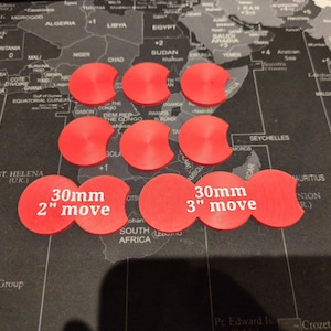K&ouml;nnte beinhalten: Neun rote Spielmarken mit gebogenem Rand sind auf einer Weltkarte angeordnet. Zwei Marken sind mit "30mm 2" move" und "30mm 3" move" beschriftet. Die Marken werden wahrscheinlich f&uuml;r Brett- oder Strategiespiele verwendet.