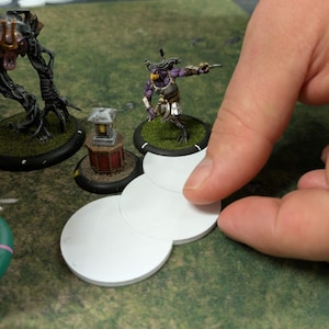 K&ouml;nnte beinhalten: Eine Tabletop-Spielszene mit Miniaturfiguren und wei&szlig;en, kreisf&ouml;rmigen Spielsteinen. Die Figuren umfassen einen lila-goldenen Krieger und ein baumartiges Wesen. Die Spielsteine werden auf einer gr&uuml;nen Oberfl&auml;che manipuliert.