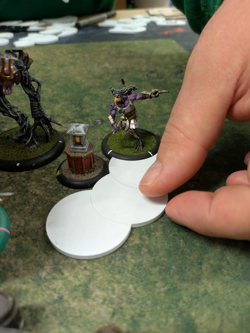 K&ouml;nnte beinhalten: Eine Tabletop-Spielszene mit Miniaturfiguren und wei&szlig;en, kreisf&ouml;rmigen Spielsteinen. Die Figuren umfassen einen lila-goldenen Krieger und ein baumartiges Wesen. Die Spielsteine werden auf einer gr&uuml;nen Oberfl&auml;che manipuliert.