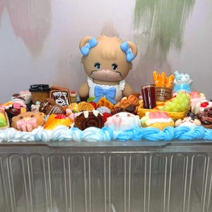 Puede incluir: Un recipiente de pl&aacute;stico transparente lleno de postres en miniatura y una figura de oso. Los postres incluyen pasteles, pasteles y otros dulces, con glaseado azul y blanco. La figura del oso tiene lazos azules y una pajarita azul.