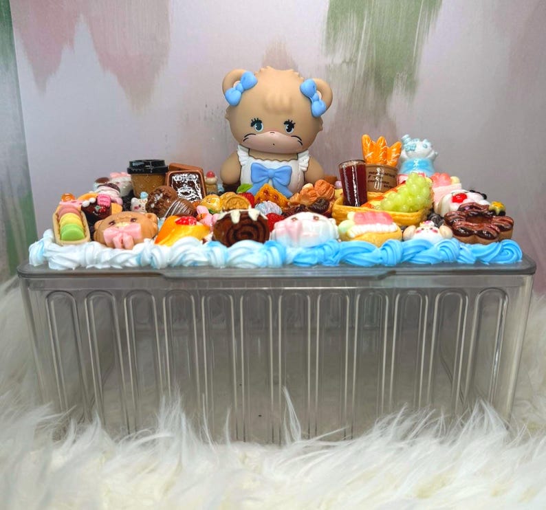 Puede incluir: Un recipiente de pl&aacute;stico transparente lleno de postres en miniatura y una figura de oso. Los postres incluyen pasteles, pasteles y otros dulces, con glaseado azul y blanco. La figura del oso tiene lazos azules y una pajarita azul.