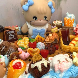 Puede incluir: Una exhibici&oacute;n caprichosa con una figura de oso con lazos azules, rodeada de miniaturas de alimentos como pasteles, fresas y un letrero de caf&eacute;. La escena es colorida y detallada, con una est&eacute;tica l&uacute;dica.