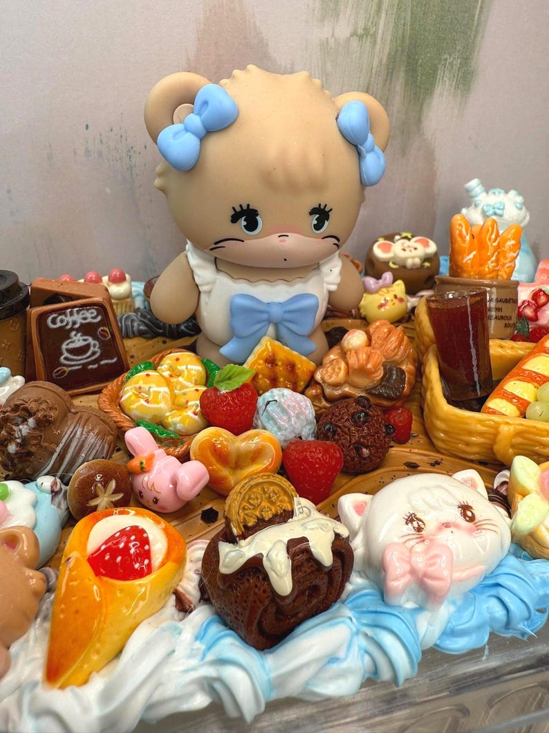 Puede incluir: Una exhibici&oacute;n caprichosa con una figura de oso con lazos azules, rodeada de miniaturas de alimentos como pasteles, fresas y un letrero de caf&eacute;. La escena es colorida y detallada, con una est&eacute;tica l&uacute;dica.