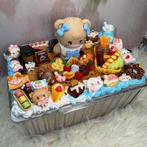 Puede incluir: Un recipiente de pl&aacute;stico transparente lleno de postres en miniatura y una figura de oso. Los postres incluyen macarons, conos de helado y pasteles. La figura del oso tiene lazos azules y un vestido blanco. El recipiente est&aacute; decorado con glaseado azul y blanco.