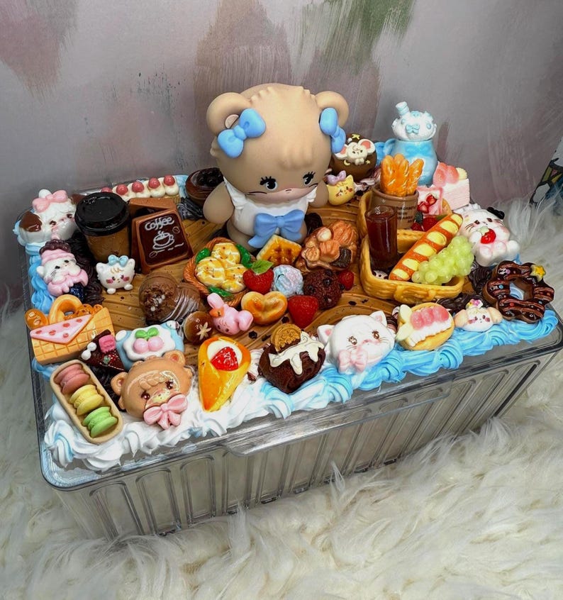 Puede incluir: Un recipiente de pl&aacute;stico transparente lleno de postres en miniatura y una figura de oso. Los postres incluyen macarons, conos de helado y pasteles. La figura del oso tiene lazos azules y un vestido blanco. El recipiente est&aacute; decorado con glaseado azul y blanco.