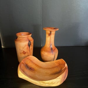 Puede incluir: Tres objetos de madera hechos a mano: un jarrón pequeño, un jarrón más alto y un cuenco curvo. La madera tiene un tono marrón rojizo cálido con vetas y nudos naturales. El cuenco tiene una base de color más claro.