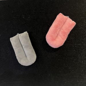 May include: Two plush, U-shaped objects. One is light gray, the other is pink. Both have a soft, fuzzy texture and a small loop at the top. The objects are set against a dark background.
