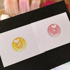 May include: Two white rectangular cards with circular designs. One design is yellow with a white heart, a cross, and a blue dot. The other is pink with a white heart, a cross, and a blue dot. The cards are on a black surface.