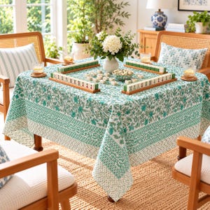 May include: A square tablecloth featuring a green and white floral block print pattern with geometric borders. The tablecloth is draped over a table set with a Mahjong game, including tiles arranged in wooden racks, a bowl of tiles, and a vase of flowers.