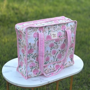 May include: A floral tote bag with pink and white flowers on a beige background. The bag has pink striped handles and trim. The text "Bloom by Miss" is visible on a small tag. The bag is sitting on a white table.