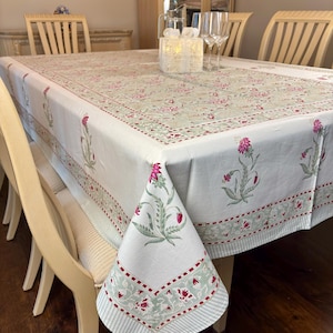 May include: A rectangular white tablecloth with a floral pattern in pink and green. The tablecloth is draped over a wooden table, with the corners hanging down. The edges have a striped border with red floral accents. The table is set with glasses and a decorative box.