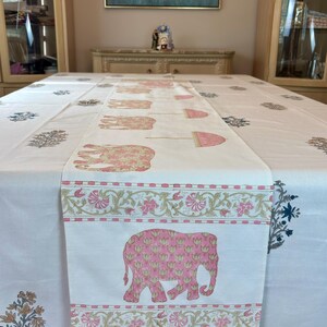 May include: White tablecloth with a pink and gold elephant and floral pattern. The table runner features a repeating elephant design and decorative borders. The tablecloth is decorated with floral and leaf patterns in various colors.
