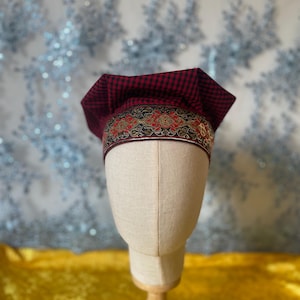 May include: A red and black checkered chef's hat with an ornate band featuring gold, red, and brown patterns. The hat is displayed on a mannequin head with a beige fabric covering. The background is a blurred pattern of blue and silver.