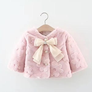May include: A light pink, plush jacket with a cream-colored bow and pearl accents. The jacket features a floral pattern and is designed with a round collar and three pearl buttons. The jacket is hanging on a wooden hanger.
