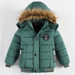 Peut inclure: Une doudoune vert foncé avec une capuche bordée de fausse fourrure marron. La veste présente un matelassage horizontal, une fermeture éclair avec boutons-pression, des poignets côtelés et un écusson circulaire avec un smiley et le texte CLASSIC FASHION 1973.