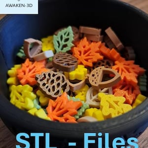 Op de afbeelding: Een zwarte kom gevuld met kleine 3D-geprinte herfstvormen, waaronder oranje esdoornbladeren, bruine dennenappels, beige eikels en gele eikenbladeren. De tekst AWAKEN-3D en STL - Files is zichtbaar in de afbeelding en duidt op digitale bestanden.