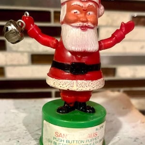 May include: A vintage Santa Claus push button puppet figurine. The red and white Santa figure has a white beard, black belt, and is holding a silver bell. The base is green with a label that reads "PUSH BUTTON PUPPET".