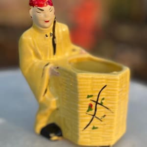 以下が含まれることがあります： 黄色いローブを着た人物の形をした、ヴィンテージのセラミックプランター。人物は赤い頭飾りと黒い髪をしています。プランターは、黒と緑のアクセントが付いたバスケットのようなデザインです。