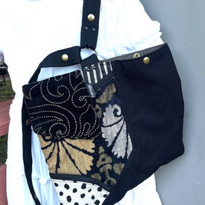 May include: A patchwork shoulder bag featuring a black canvas side, a black velvet panel with white swirling patterns, a gold and black floral textile section, and a white triangular base with black polka dots. It has a black strap with brass rivets.
