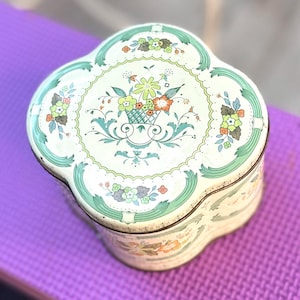 Puede incluir: Una lata de metal con forma de trébol y base color crema, que presenta una ilustración central de una cesta de flores en tonos verdes, naranjas y marrones. La tapa está decorada con intrincados adornos verdes y flores.