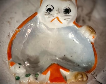 Può includere: Un piatto a forma di gatto in ceramica con corpo bianco, dettagli arancioni e interno azzurro. Il gatto ha grandi occhi neri, un piccolo naso arancione e baffi. Una piccola statuina di gatto è nel piatto. L'oggetto è decorativo.