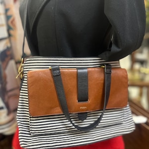 May include: A Fossil crossbody bag with a brown leather flap and black strap closure. The bag features a black and white striped pattern and a brown leather front. The Fossil logo is visible on the front.