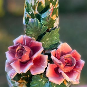 Peut inclure: Un vase en céramique avec une base crème et un sommet évasé. Le vase est orné de motifs floraux peints à la main et présente deux grandes roses roses avec des feuilles vertes. Le vase mesure environ 30 cm de haut.